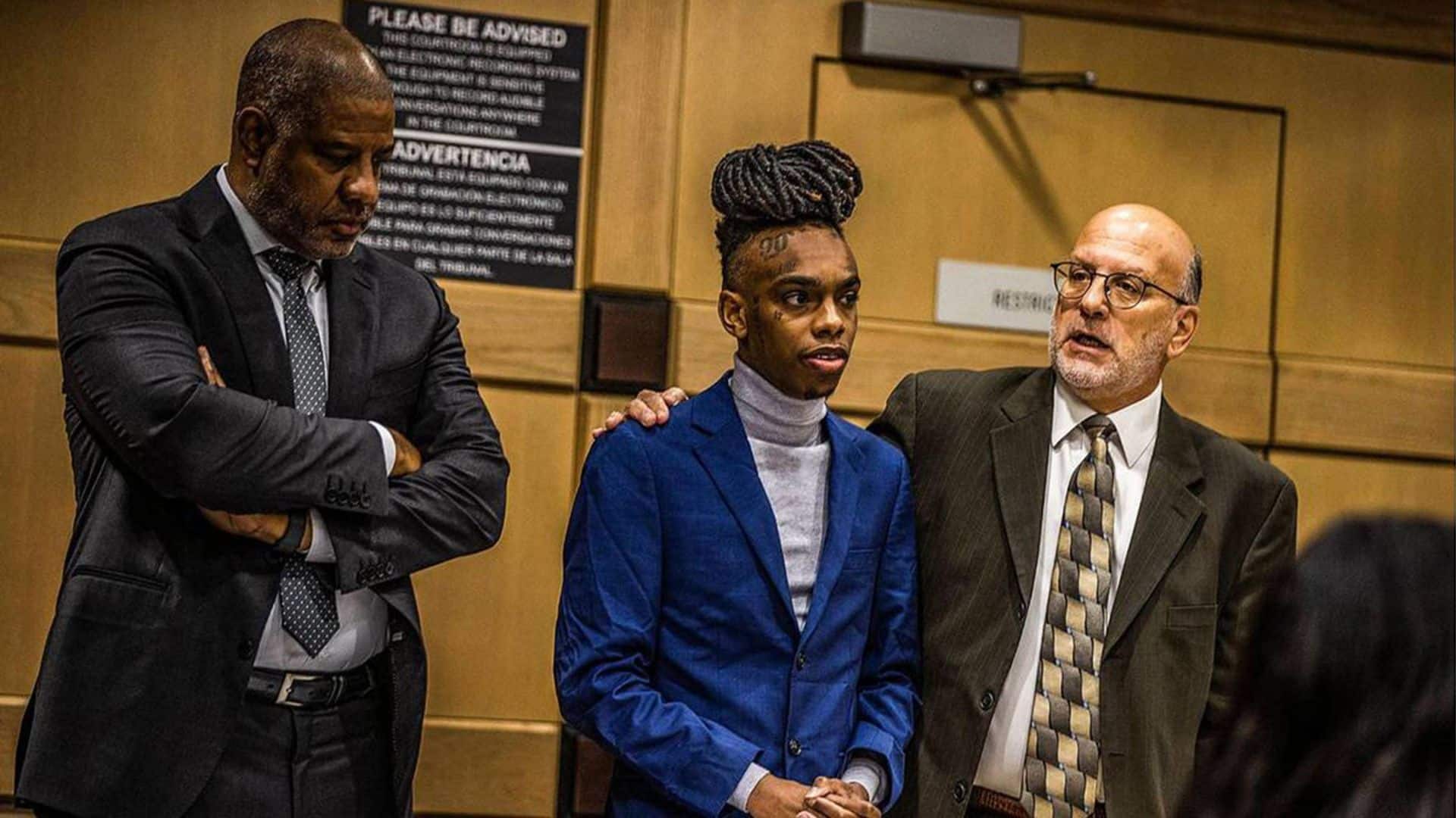 YNW Melly in a blue suit standing in a courtroom with two attorneys beside him.