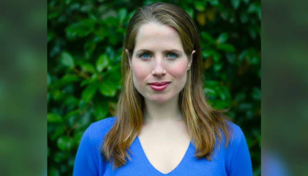 a portrait of kristin gore wearing a blue sweater with a neutral expression standing against a green background