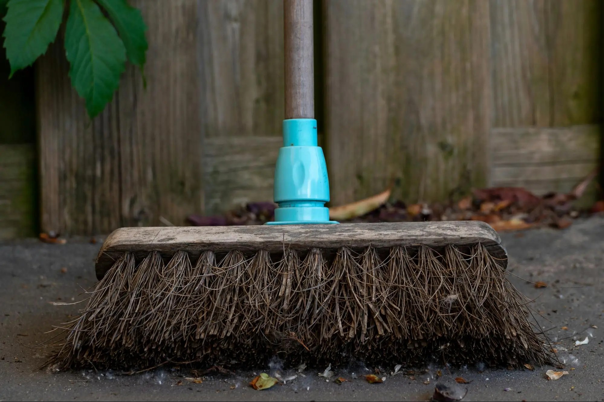 Wooden broom with blue handle on concrete pavement against wooden fence background