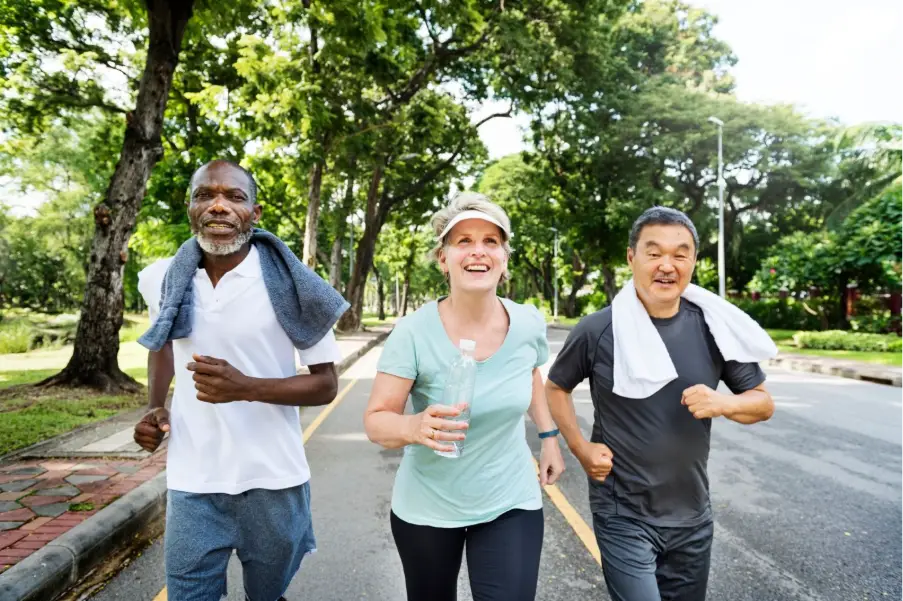Three people jogging on a tree-lined road in a park with towels and water bottle