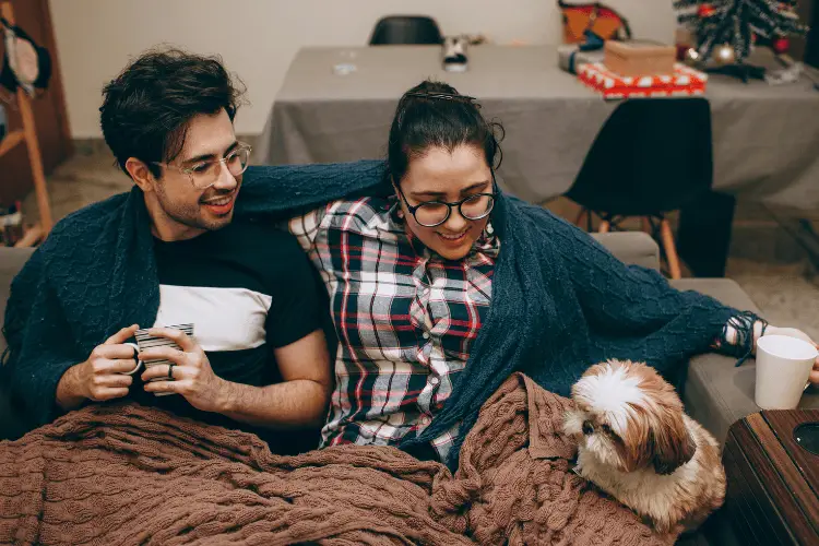Cozy couple with drinks sitting on couch under blanket, small dog beside them