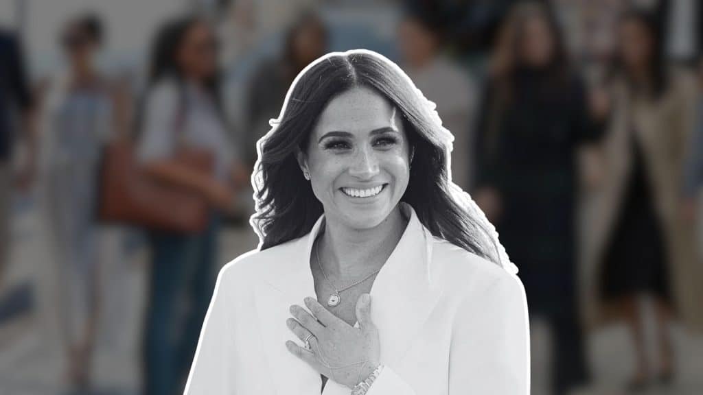 Meghan Markle smiling in a white outfit, hand over her chest, against a blurred crowd background.