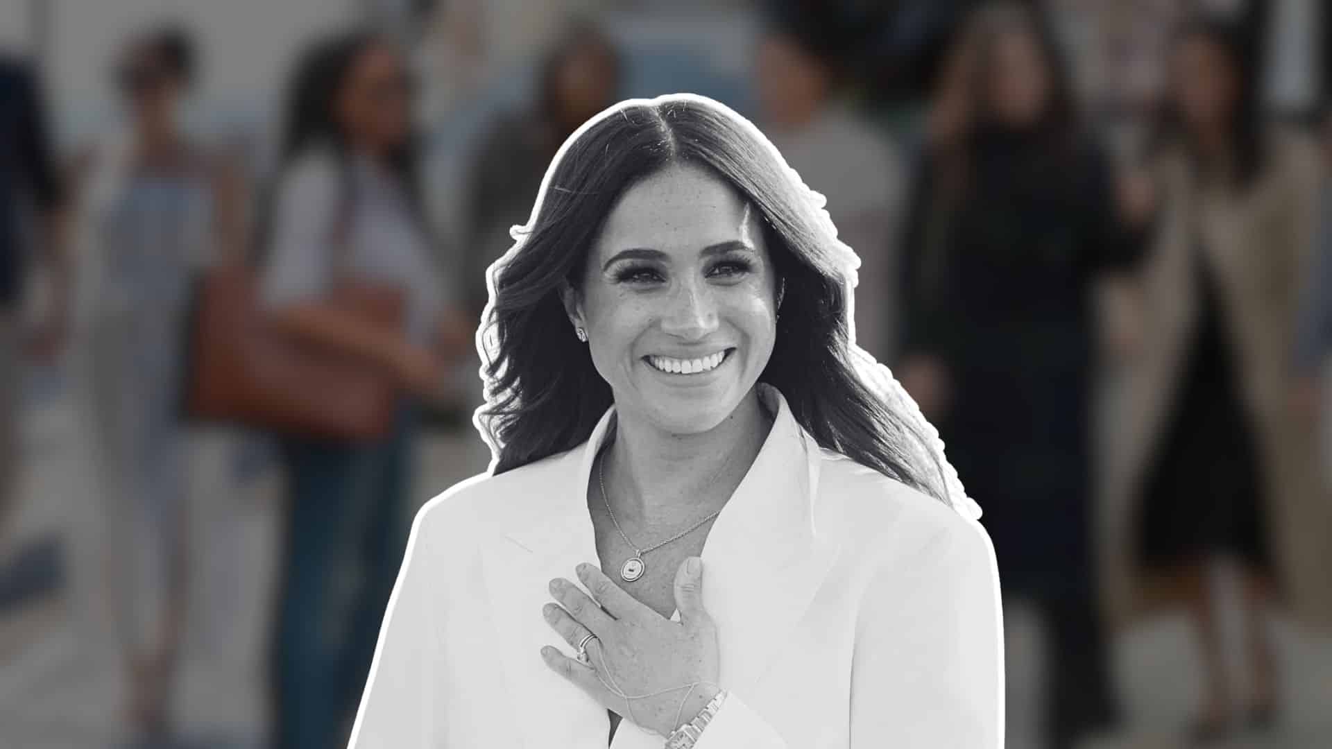 Meghan Markle smiling in a white outfit, hand over her chest, against a blurred crowd background.