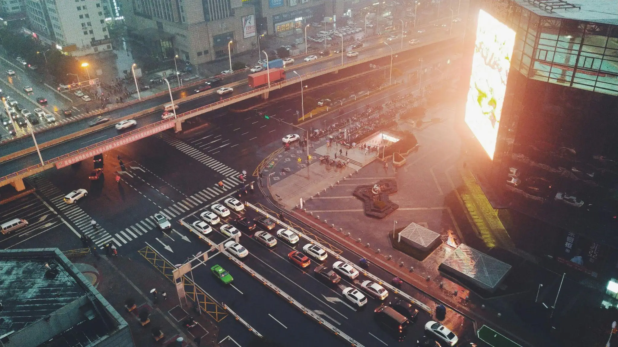 Busy urban intersection with cars and pedestrians, large digital billboard at dusk in cityscape