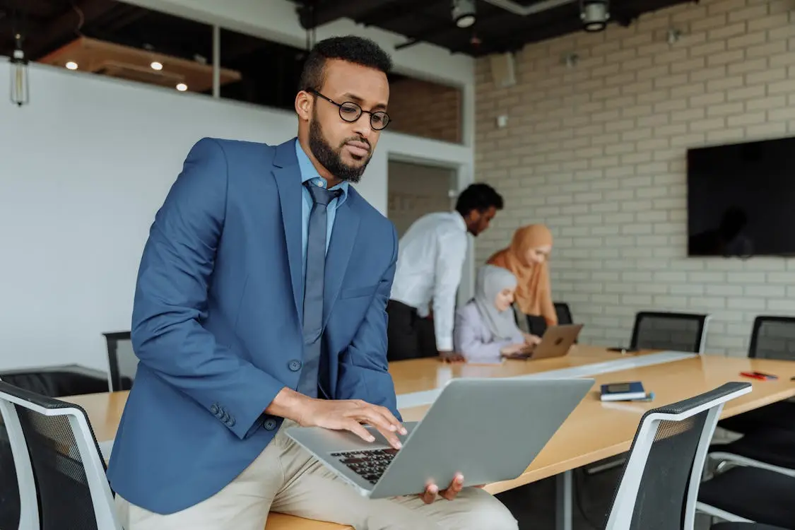 Man in blue suit using laptop in modern conference room with colleagues in background