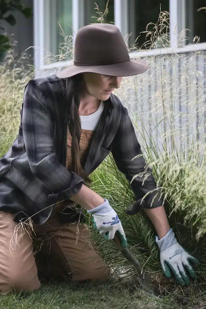 Gardener wearing brown hat and plaid shirt tending tall grass with hand spade