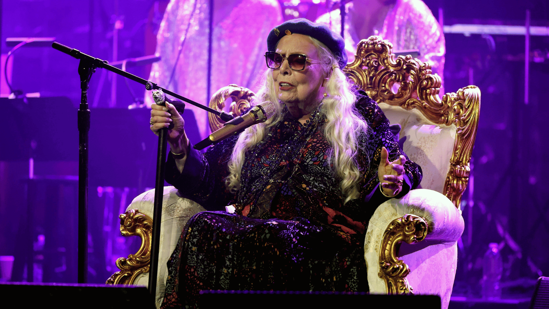 Joni Mitchell performing live on stage under purple lighting while seated in ornate chair.