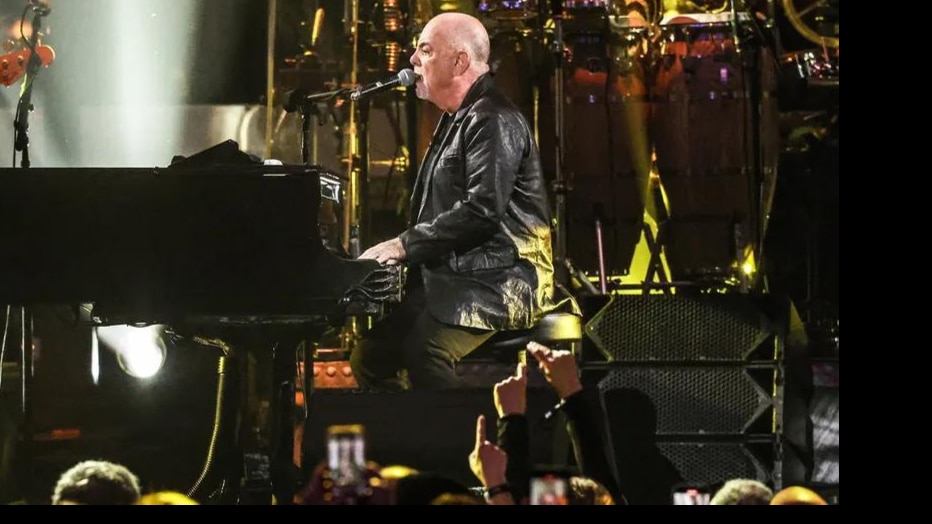 Billy Joel performing live in concert, singing at the piano in front of a packed crowd during an energetic arena show.