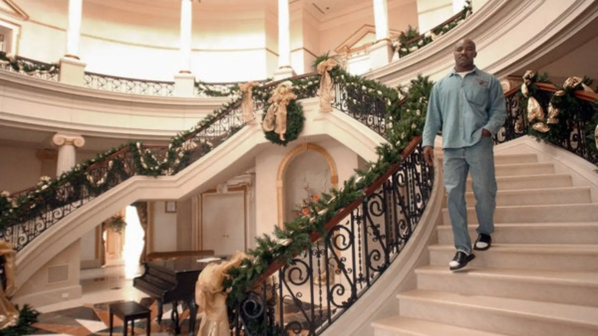 Evander Holyfield walks down the grand staircase inside his lavish Fayetteville mansion, featuring ornate railings, towering columns, and elegant holiday decorations