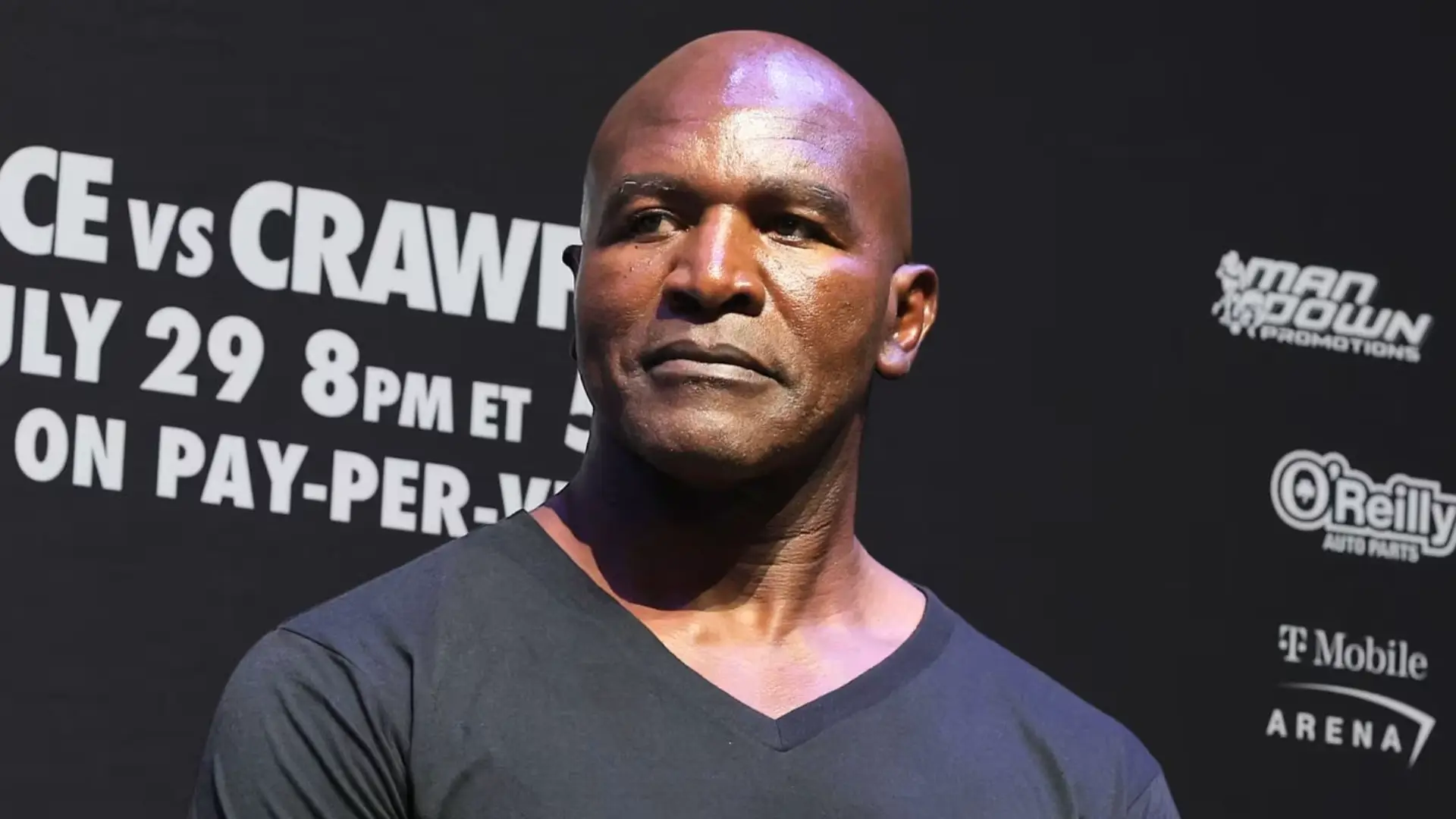 Evander Holyfield standing at a press event, looking to the side with a neutral expression, wearing a black shirt in front of a promotional backdrop