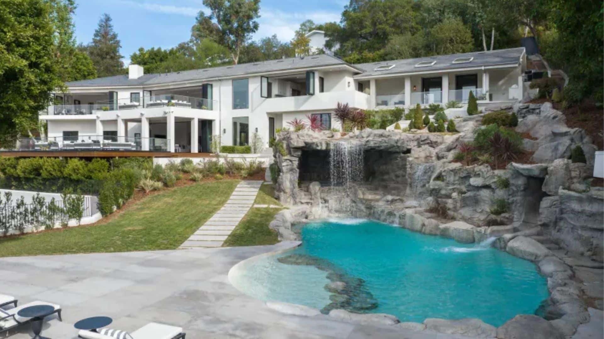 Mediterranean-style Beverly Hills estate on Oak Pass Road, featuring a large main house with multiple wings, a two-story gym, grotto-style pool with waterfall, lush gardens, and outdoor sports areas under a clear sky.