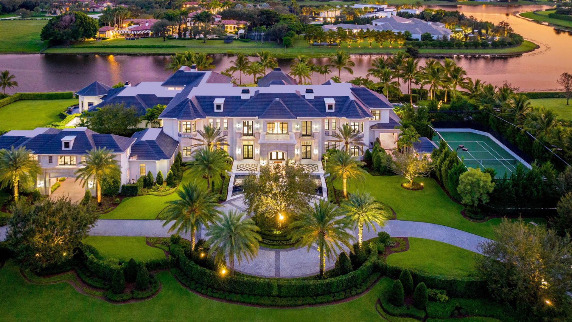 Lakefront luxury mansion Palazzo di Lago in Delray Beach, Florida, with a lagoon-style pool, waterfall, fire features, and lush landscaping under a clear sky.