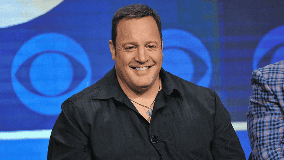 Smiling Kevin James at a panel event in a black shirt, representing the star behind the massive kevin james net worth.