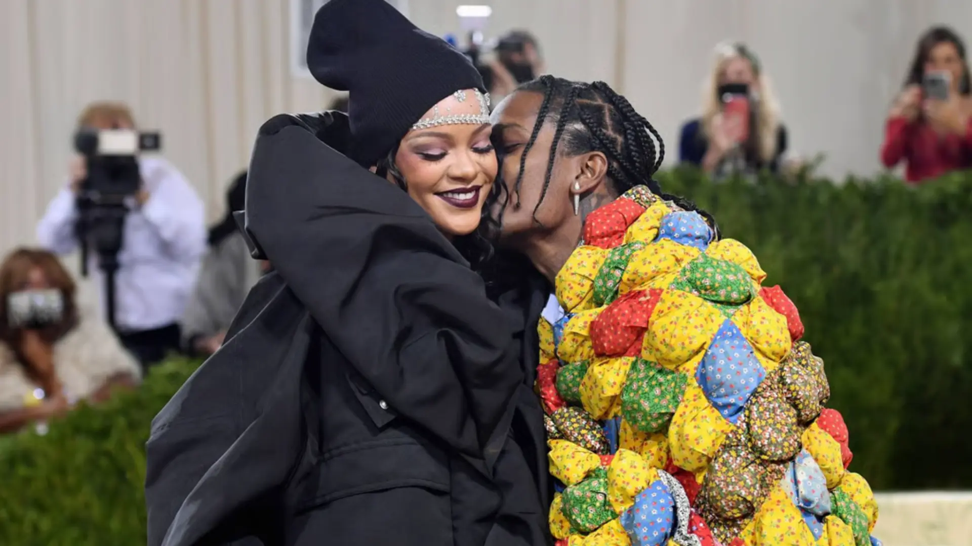 Rihanna and a$sap rocky at met gala event, one wearing a black structured outfit and headpiece smiling, while another in a colorful patchwork outfit