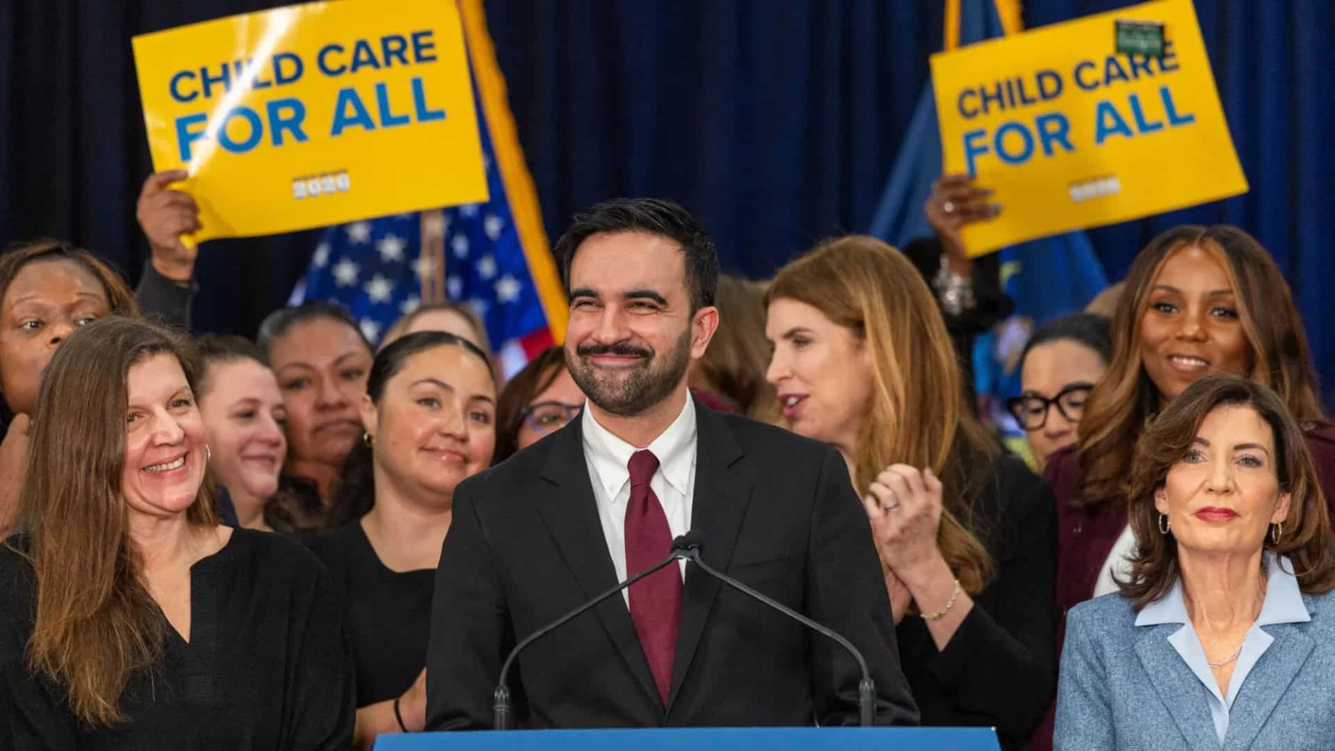 Zohran Mamdani stands at a podium delivering a speech with a group of supporters behind him. Signs in the background read “CHILD CARE FOR ALL,” and the podium displays a message about family and future priorities.