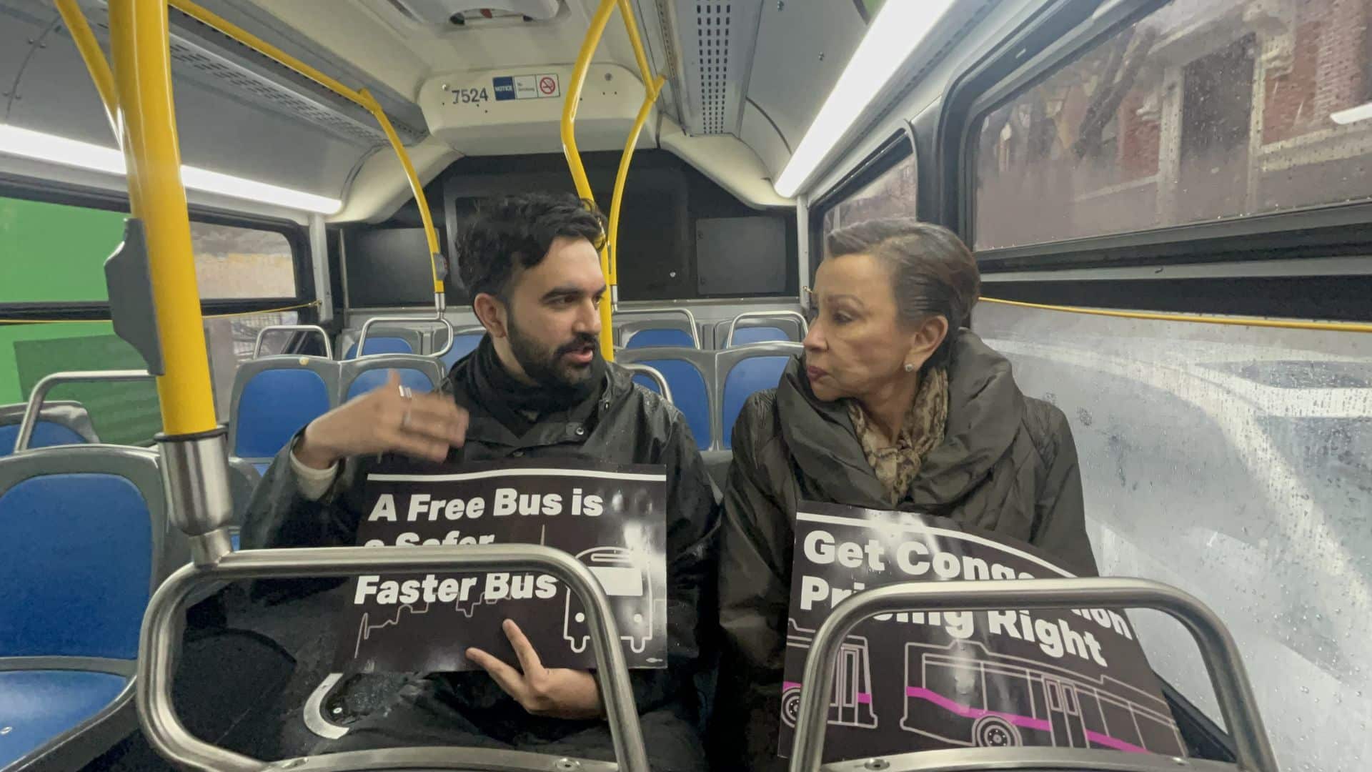Zohran Mamdani sits on a city bus holding a sign advocating for free and faster buses, speaking with a woman next to him who also holds a campaign sign.