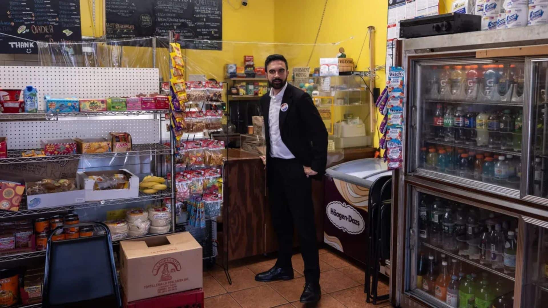 Zohran Mamdani stands inside a small grocery store, surrounded by shelves of snacks, drinks, and packaged goods. He is wearing a suit and looking toward the camera, highlighting a local business setting.