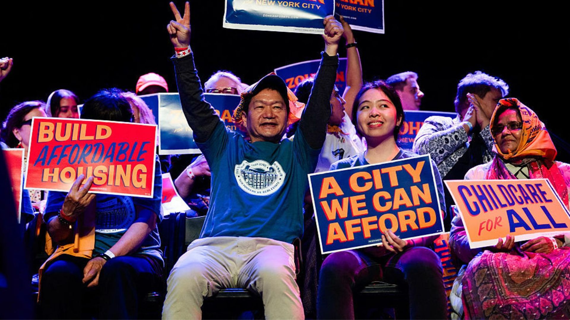 Supporters at a rally hold signs backing Zohran Mamdani, including messages on affordable housing and childcare, with one person raising a peace sign.