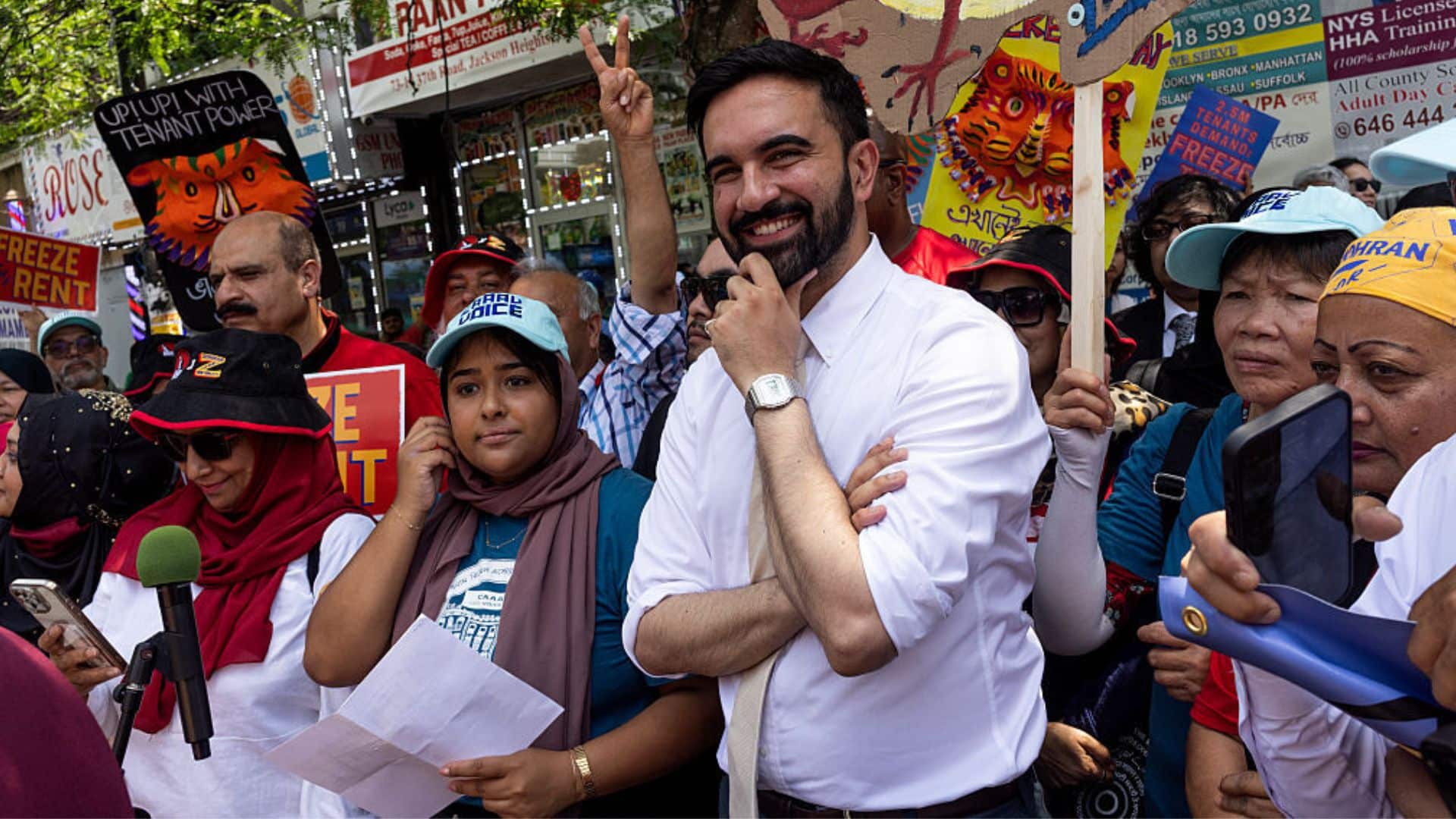 Zohran Mamdani stands smiling in a crowd of supporters at an outdoor rally. People around him hold signs advocating for rent freezes and tenant rights, and some are taking photos or reading prepared statements.
