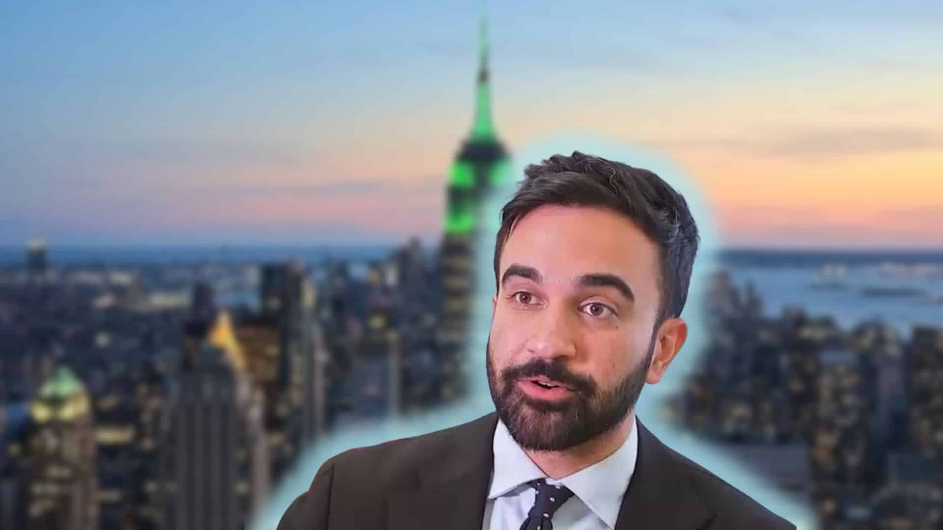 Zohran Mamdani in a dark suit and tie with a beard, speaking in front of a blurred cityscape at sunset featuring the Empire State Building lit in green.
