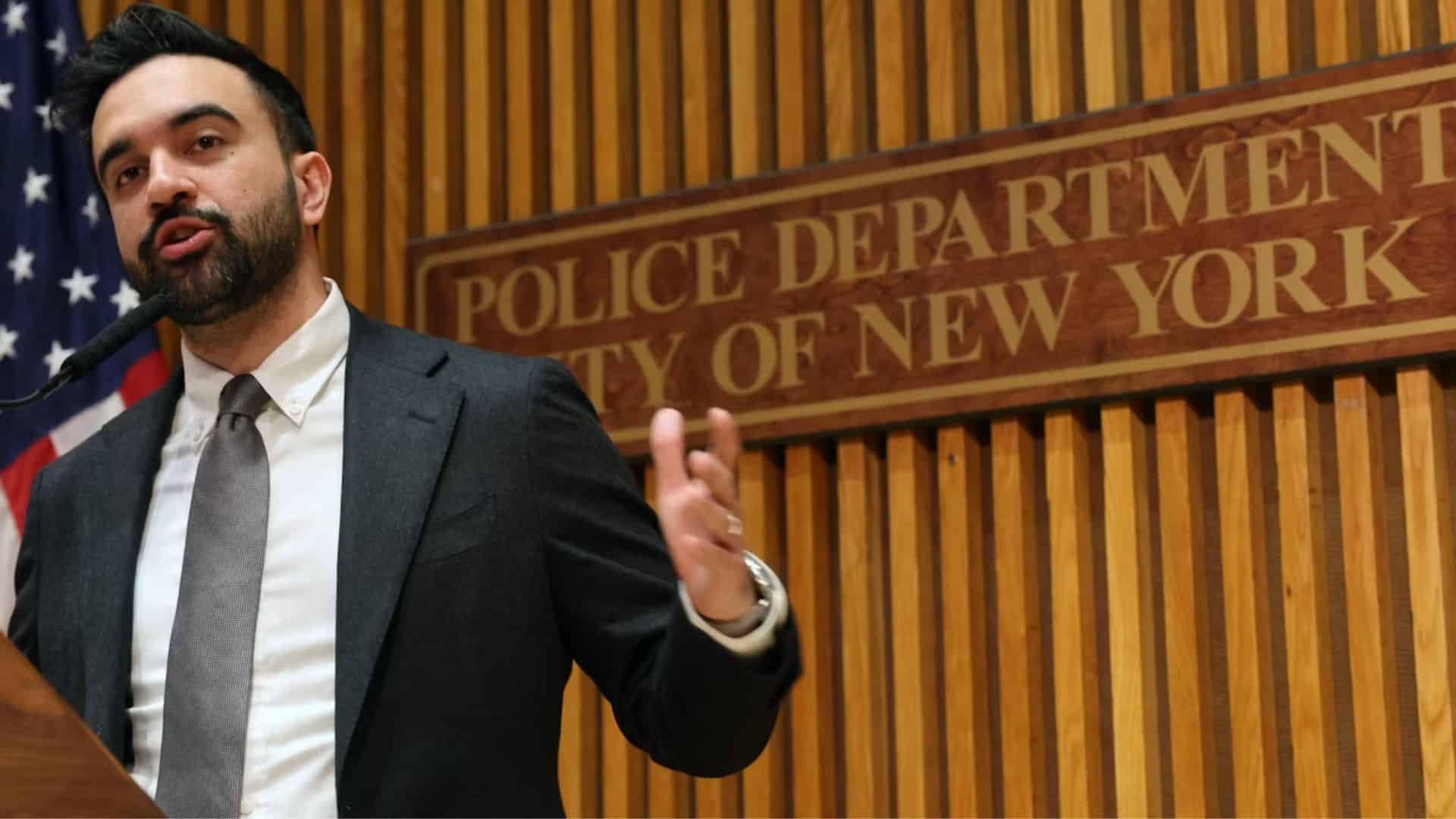 Zohran Mamdani stands at a podium with the New York Police Department emblem, speaking to the press about policing policies.