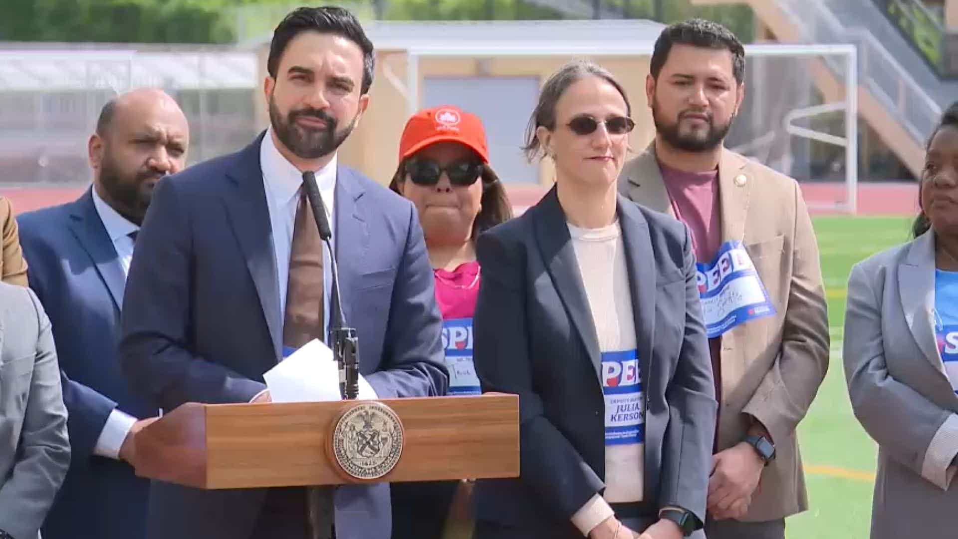 Zohran Mamdani stands at a podium speaking outdoors, surrounded by colleagues and supporters. They are dressed in professional attire, some wearing campaign shirts, listening attentively during a public event.