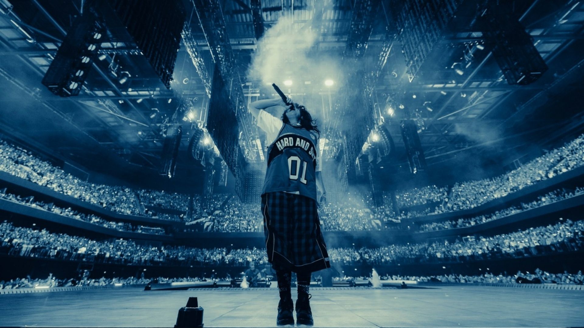 Billie Eilish performing live on a large stadium stage under blue lighting and smoke effects