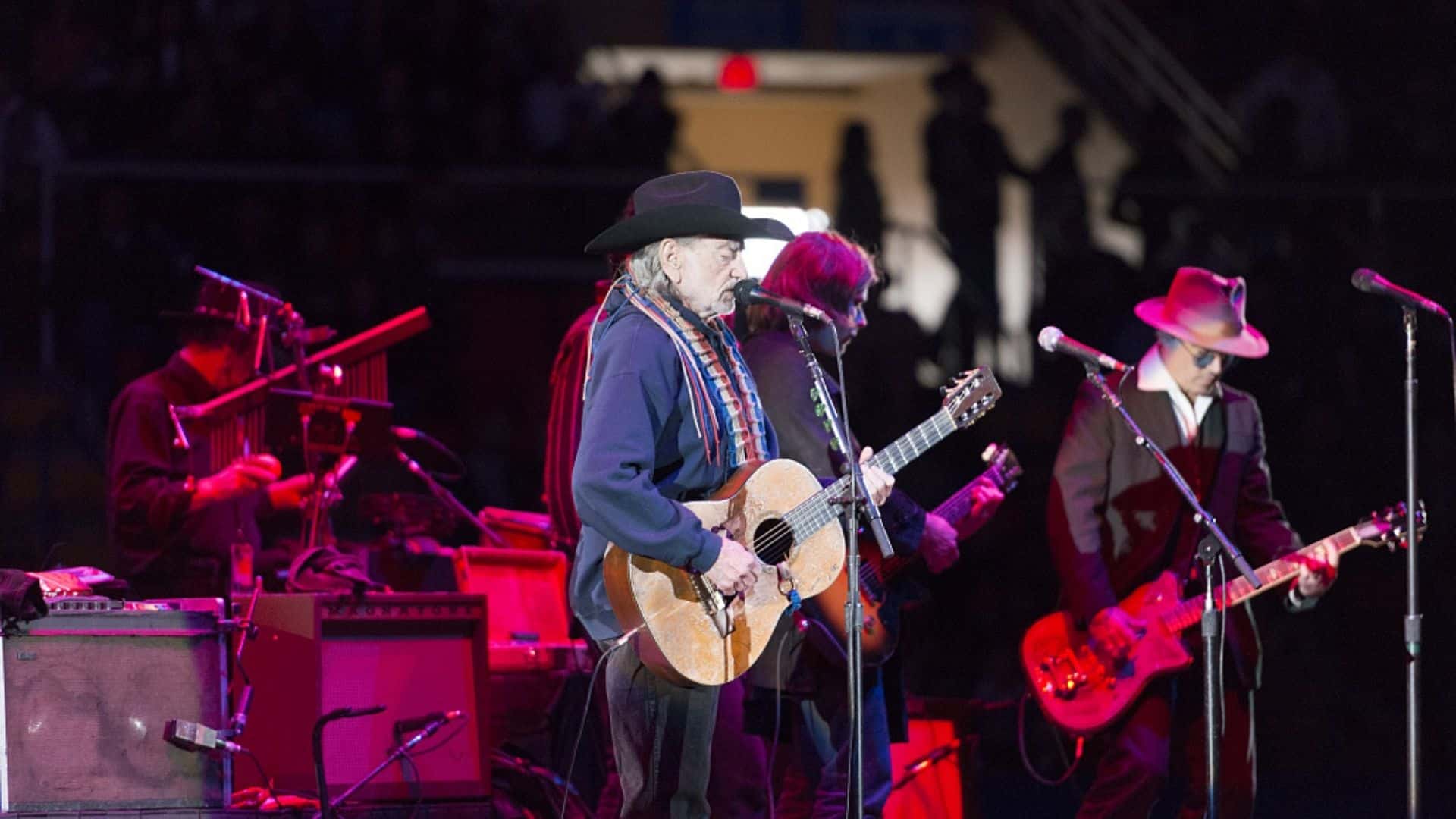 Willie Nelson performing live on stage with band members playing guitars under red stage lighting