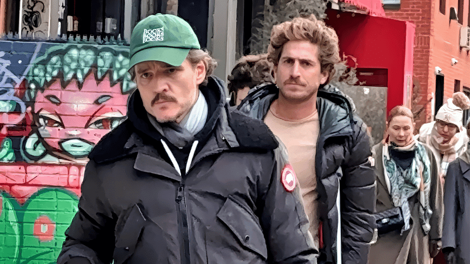 pedro pascal in green cap and dark jacket walking past graffiti wall on a busy urban street with people in background.