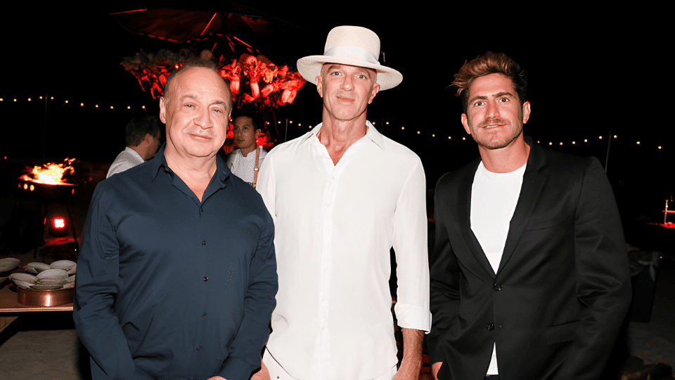 Rafael Olarra standing in a black suit beside two men at a night event with warm lights and festive background.