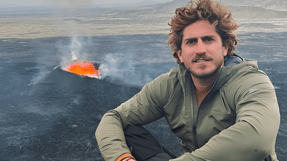 Rafael Olarra sitting outdoors on rocky terrain near a smoky wildfire in the background, looking toward the camera.