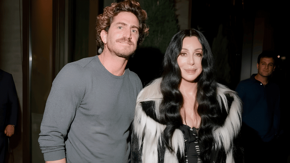 Man in a grey sweater standing beside a woman in a black outfit with fur accents, posing outdoors at night with security in background.