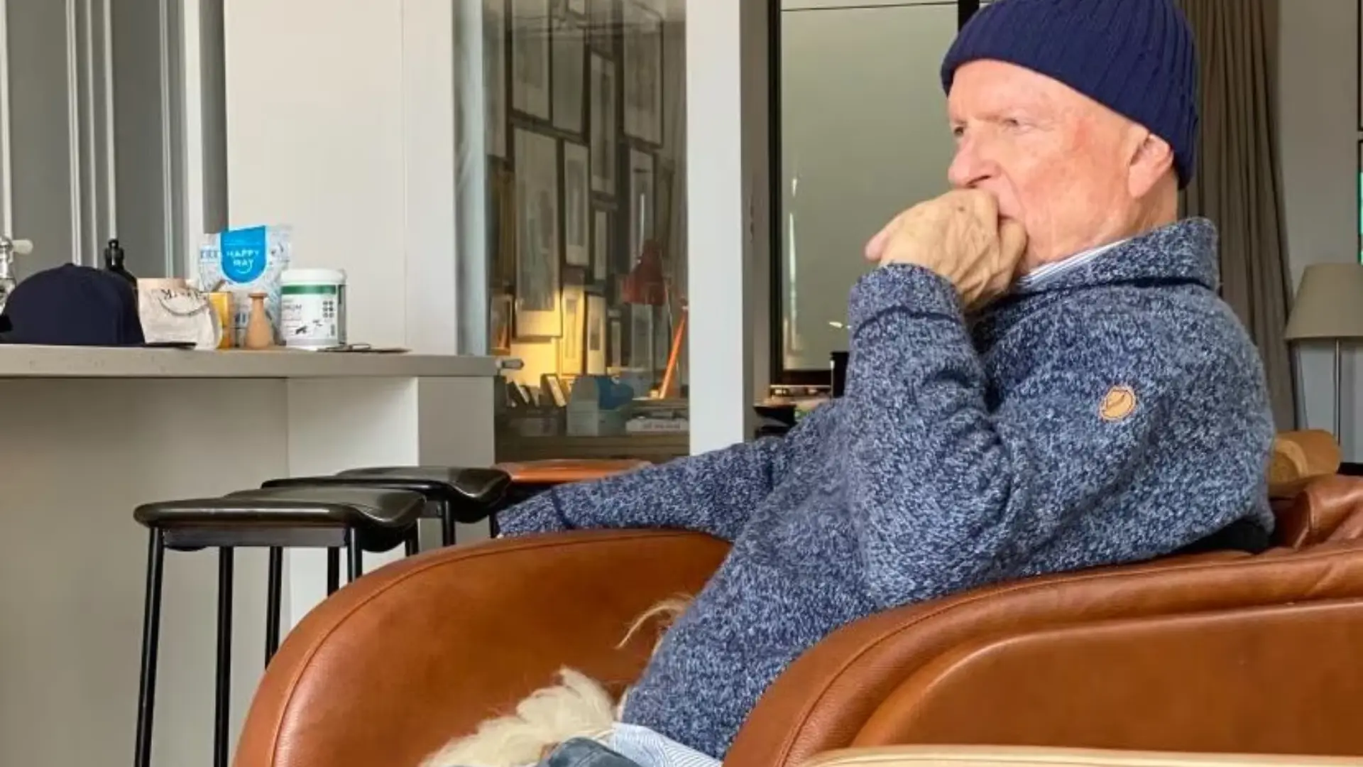 Sam Neill sitting indoors wearing a blue sweater and beanie in a casual setting.