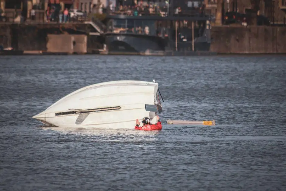 Capsized boat in river with person in life jacket near city shoreline